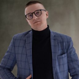 Mateusz Sidor - Doradca Ubezpieczeniowy - Mężczyzna w okularach i niebieskiej marynarce w kratę, czarny golf, pozuje na rozmowie kwalifikacyjnej, stonowane tło, profesjonalny wizerunek.