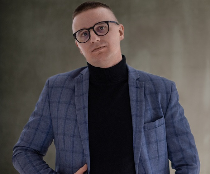 Mężczyzna w okularach i niebieskiej marynarce w kratę, czarny golf, pozuje na rozmowie kwalifikacyjnej, stonowane tło, profesjonalny wizerunek.