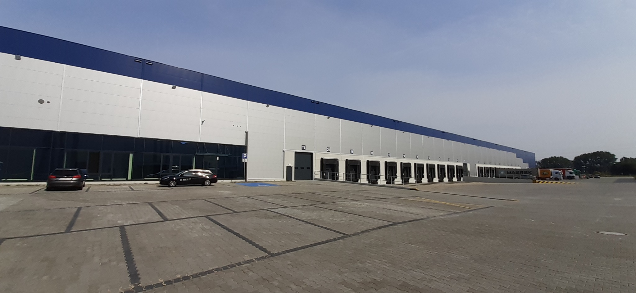 Szeroki kadr na nowoczesny magazyn z rampami załadunkowymi i szarym parkingiem z dwoma samochodami osobowymi oraz zaparkowanymi ciężarówkami w oddali.