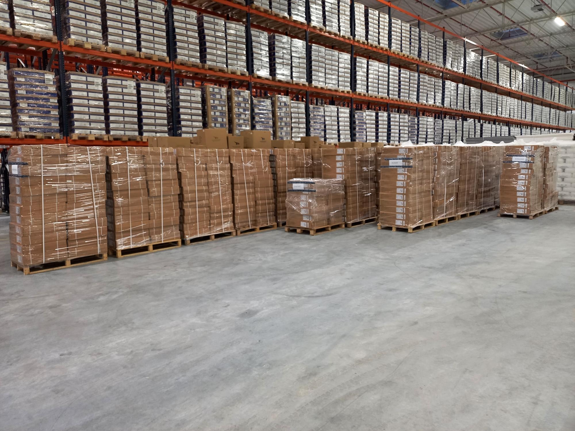 Palety z kartonami owinięte folią stretch na betonowej posadzce magazynu, regały magazynowe z towarem w tle.