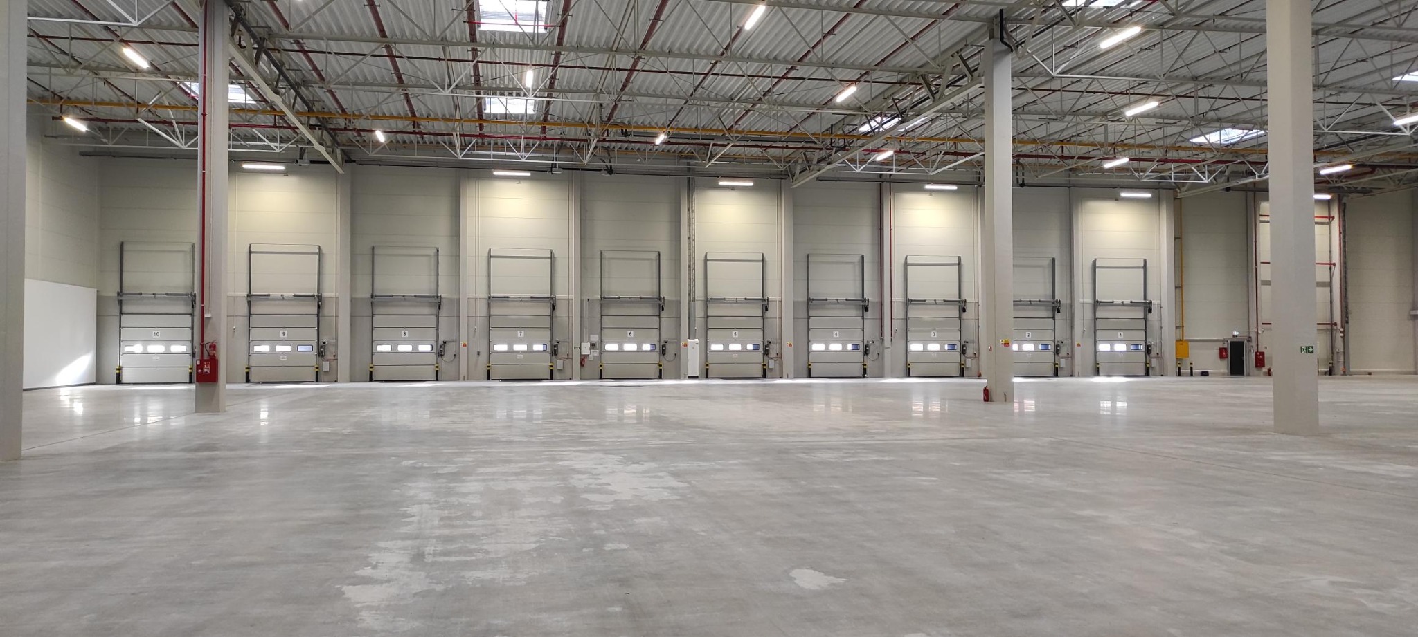 Wnętrze pustego, przestronnego magazynu z betonową posadzką, rzędem bram segmentowych z numerami i oświetleniem LED pod sufitem