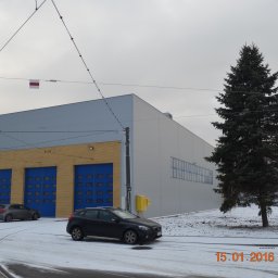 Zakład Ogólnobudowlany Bogdan Pociejewski - Duży budynek z żółtej cegły z niebieskimi bramami garażowymi, przylegający do budynku z jasnoszarej blachy trapezowej, tramwajowe tory i samochody na ośnieżonej drodze, flaga na przewodach...