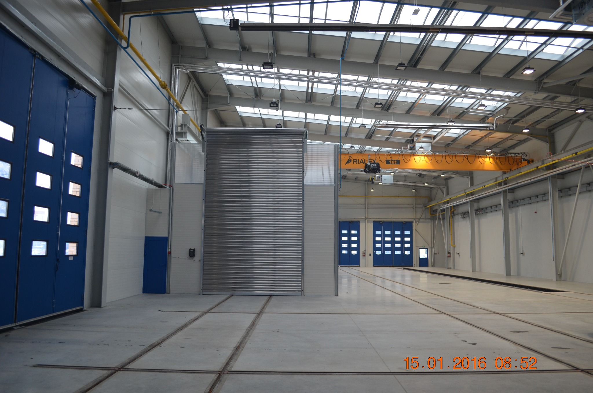 Wnętrze hali produkcyjnej z betonową posadzką, niebieskimi drzwiami segmentowymi z okienkami i metalową bramą rolowaną, widoczny suwnica pod stropem