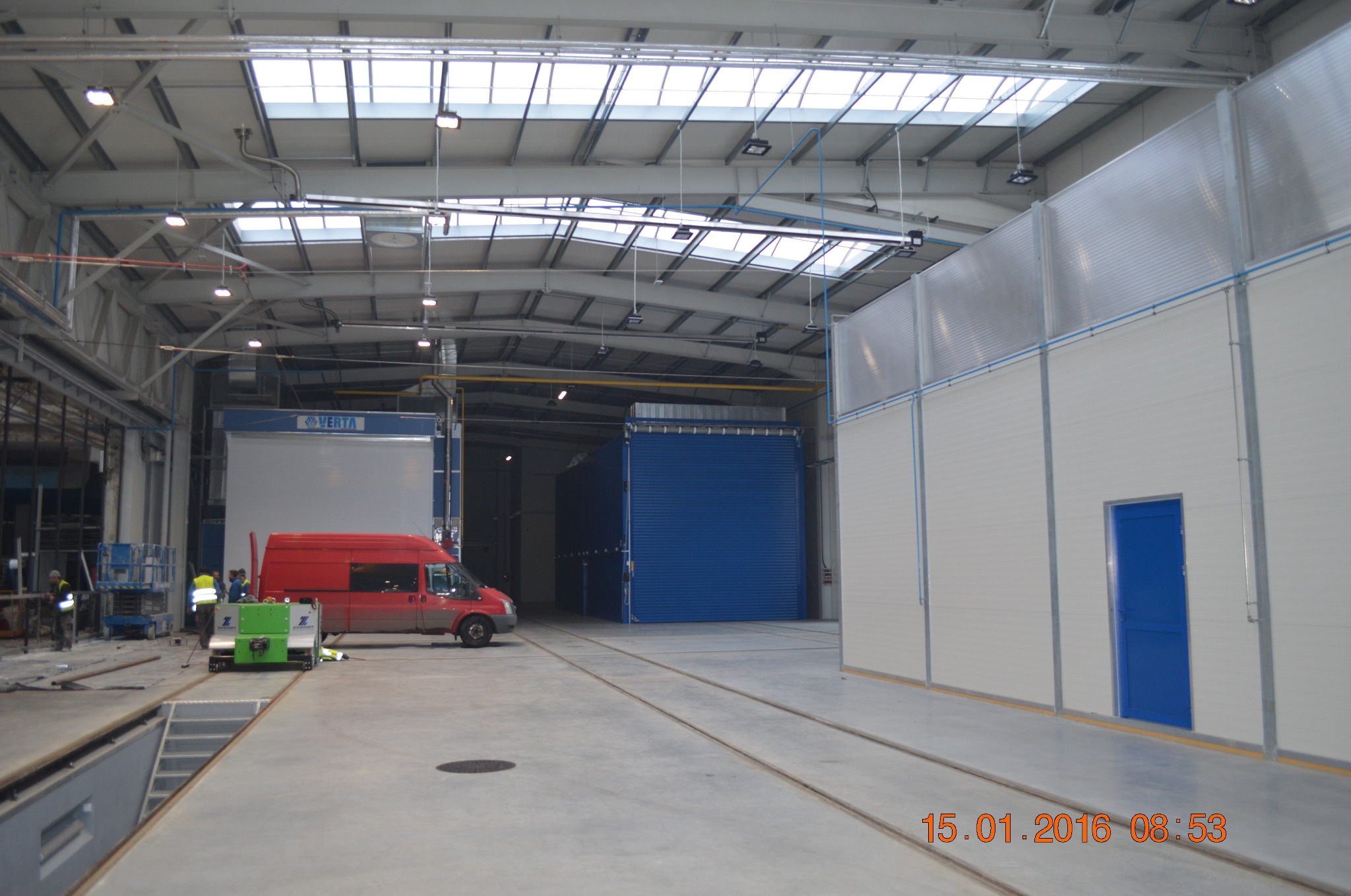 Wnętrze hali przemysłowej z oknami dachowymi, widoczne panele ścienne, brama wjazdowa, czerwony bus i pracownicy w odblaskowych kamizelkach, torowisko w posadzce.