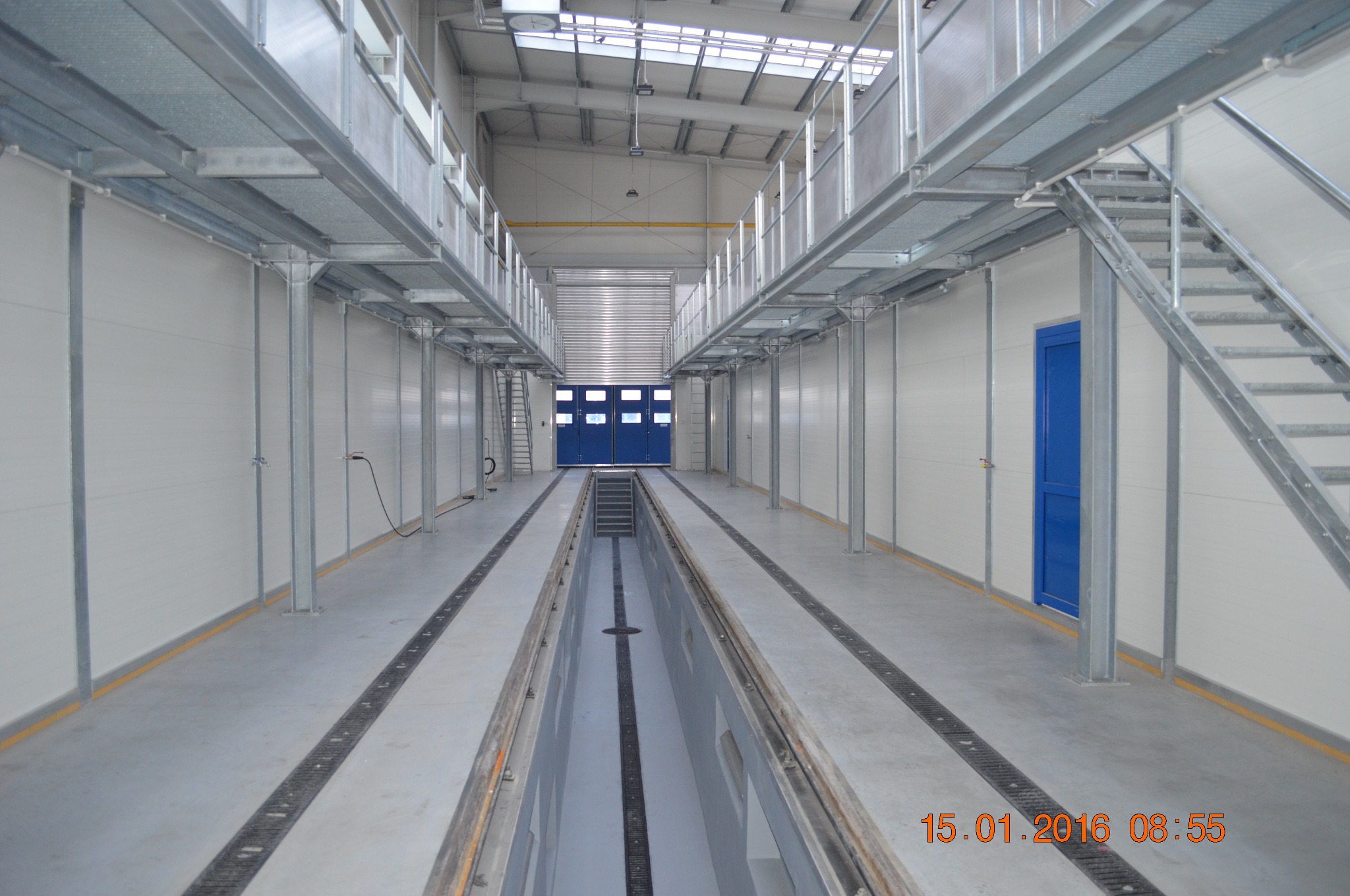 Wnętrze hali przemysłowej z kanałem rewizyjnym, widoczne białe ściany z panelami, metalowe konstrukcje pod sufitem, niebieskie drzwi i schody, w tle brama wjazdowa.