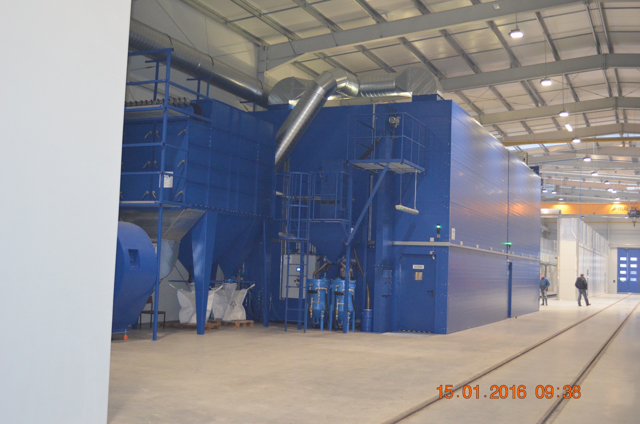 Duża, niebieska maszyna przemysłowa z systemem rur w hali fabrycznej, z widocznymi torami w podłodze i pracownikami w tle.