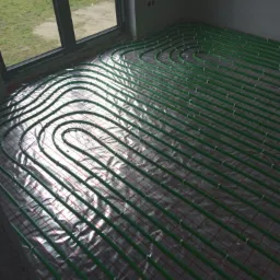 Rozłożone zielone rury ogrzewania podłogowego na folii z czerwoną siatką w surowym wnętrzu, widok z góry, światło dzienne z okna.