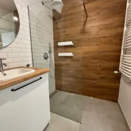 Nowoczesna łazienka po remoncie w Świdnicy: kabina prysznicowa typu walk-in z drewnopodobnymi płytkami, umywalka z blatem z jasnego drewna i białą szafką, białe płytki na ścianie. Głowica...