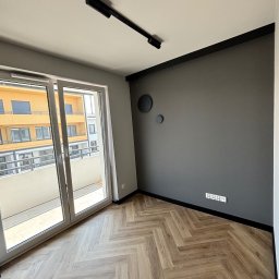 Lukbud - Nowoczesne wnętrze: szara ściana z dekorami, parkiet w jodełkę, przesuwne drzwi balkonowe z widokiem na budynek. Minimalistyczne oświetlenie sufitowe.