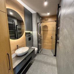 Lukbud - Nowoczesna łazienka z drewnianymi i szarymi akcentami. Umywalka nablatowa, okrągłe lustro z podświetleniem LED i kabina prysznicowa typu walk-in.
