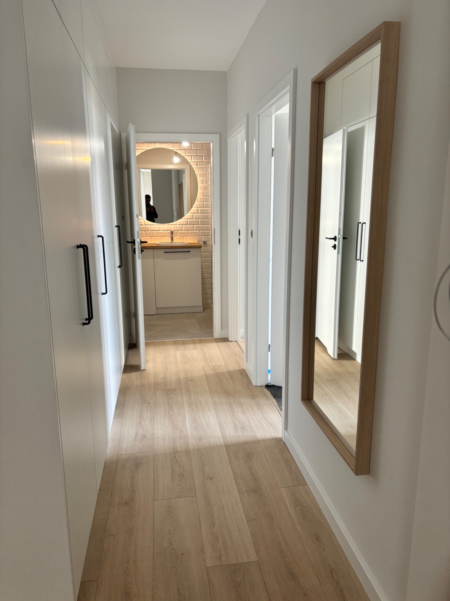 Długi korytarz z zabudową szaf w kolorze białym, na podłodze panele w odcieniach beżu, w głębi widoczna otwarta łazienka z okrągłym lustrem i beżowymi płytkami.