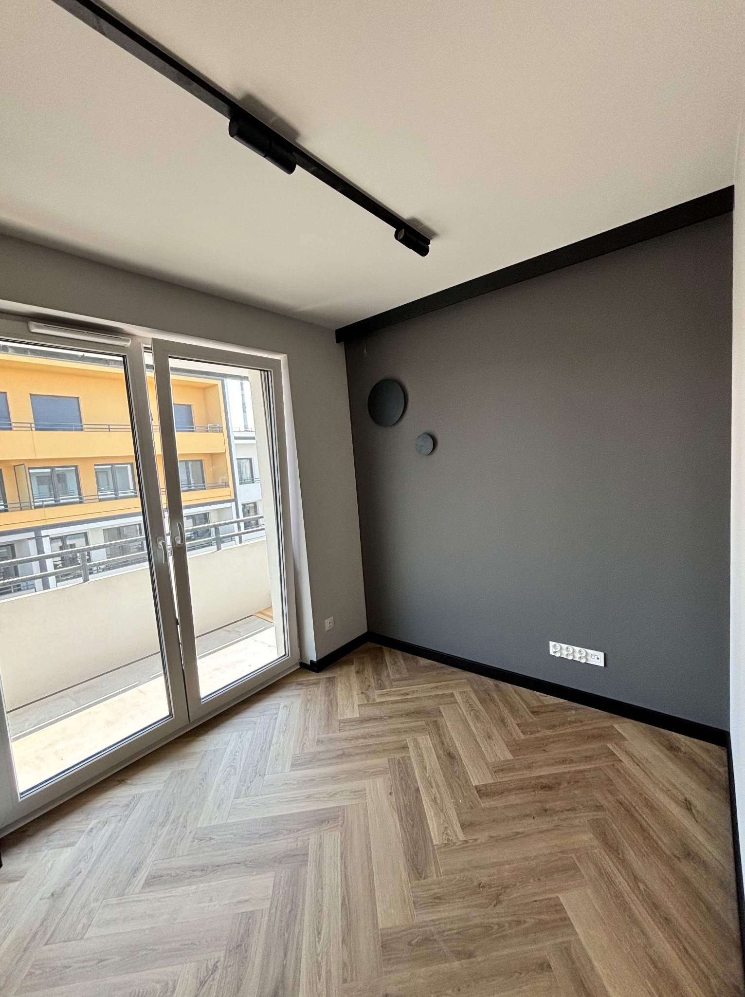 Nowoczesne wnętrze: szara ściana z dekorami, parkiet w jodełkę, przesuwne drzwi balkonowe z widokiem na budynek. Minimalistyczne oświetlenie sufitowe.