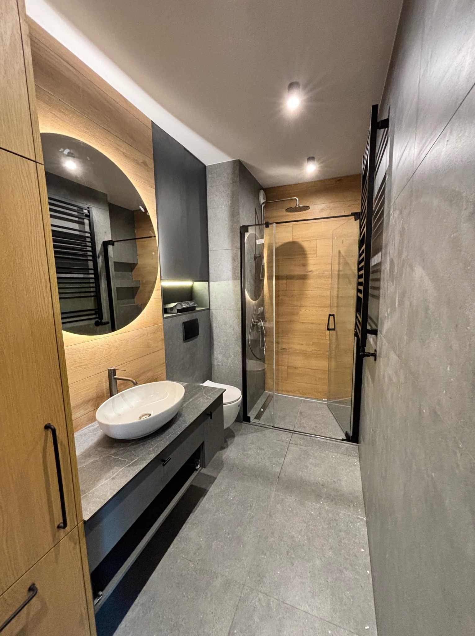Nowoczesna łazienka z drewnianymi i szarymi akcentami. Umywalka nablatowa, okrągłe lustro z podświetleniem LED i kabina prysznicowa typu walk-in.