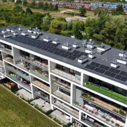 Nowoczesny apartamentowiec z panelami fotowoltaicznymi na dachu, widok z góry, balkony z roślinami, w tle zieleń i tory kolejowe.
