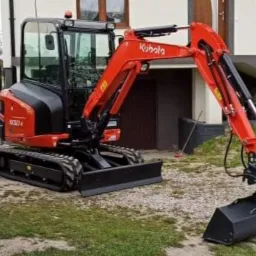 Czerwono-czarna mini koparka Kubota na żwirowym podłożu, z łyżką skierowaną w dół, zaparkowana przed budynkiem z widocznymi schodami.