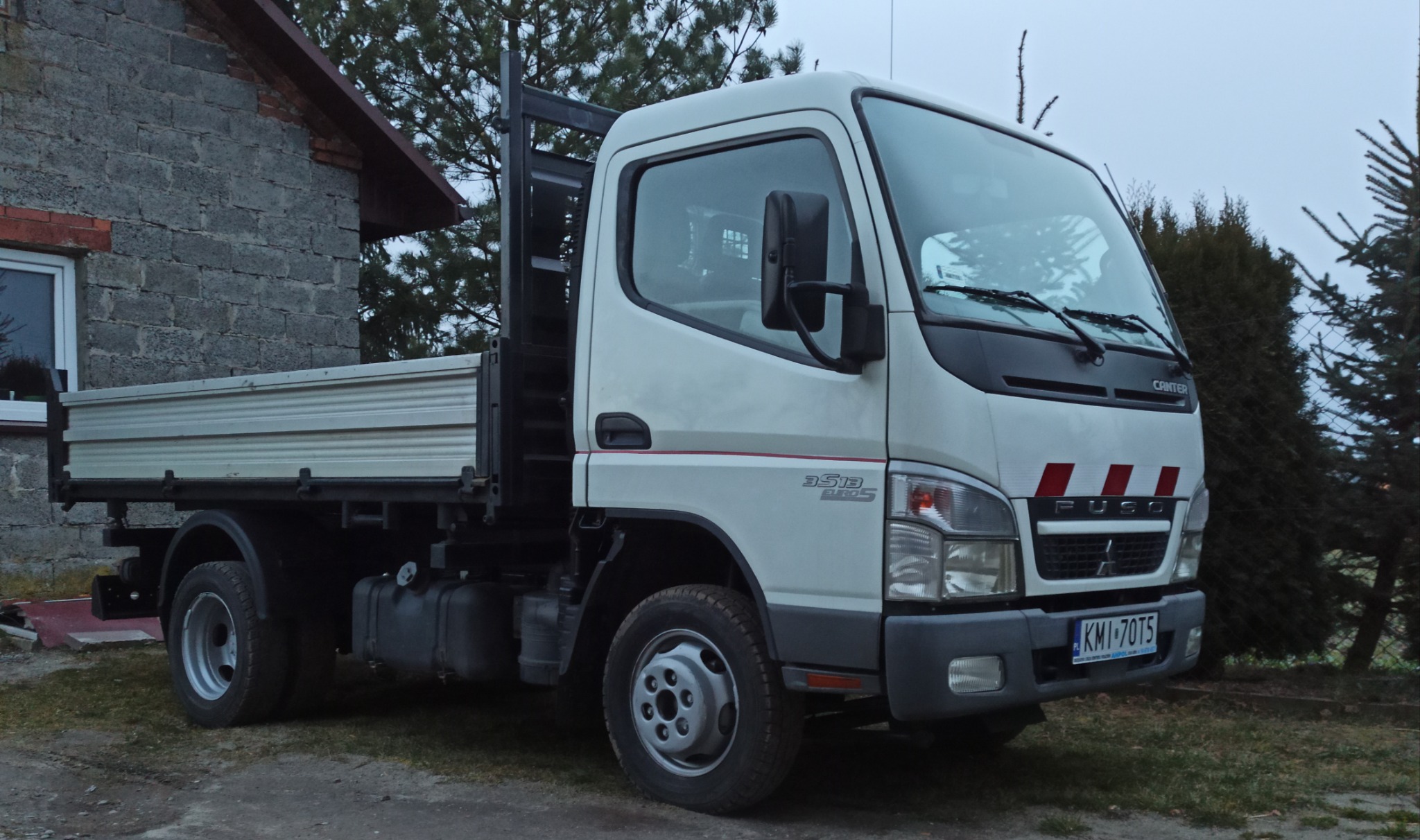 Biała ciężarówka Fuso Canter 3.5t z otwartą skrzynią ładunkową, zaparkowana obok szarego budynku z bloczków betonowych i zielonych drzew iglastych.