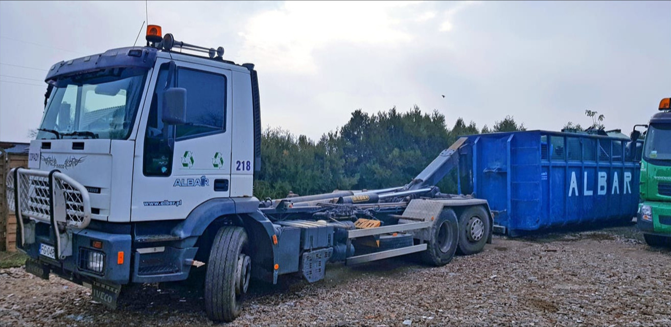 Ciężarówka z hakiem na podwoziu Iveco z niebieskim kontenerem na gruz firmy Albar, na żwirowym placu, w tle drzewa.