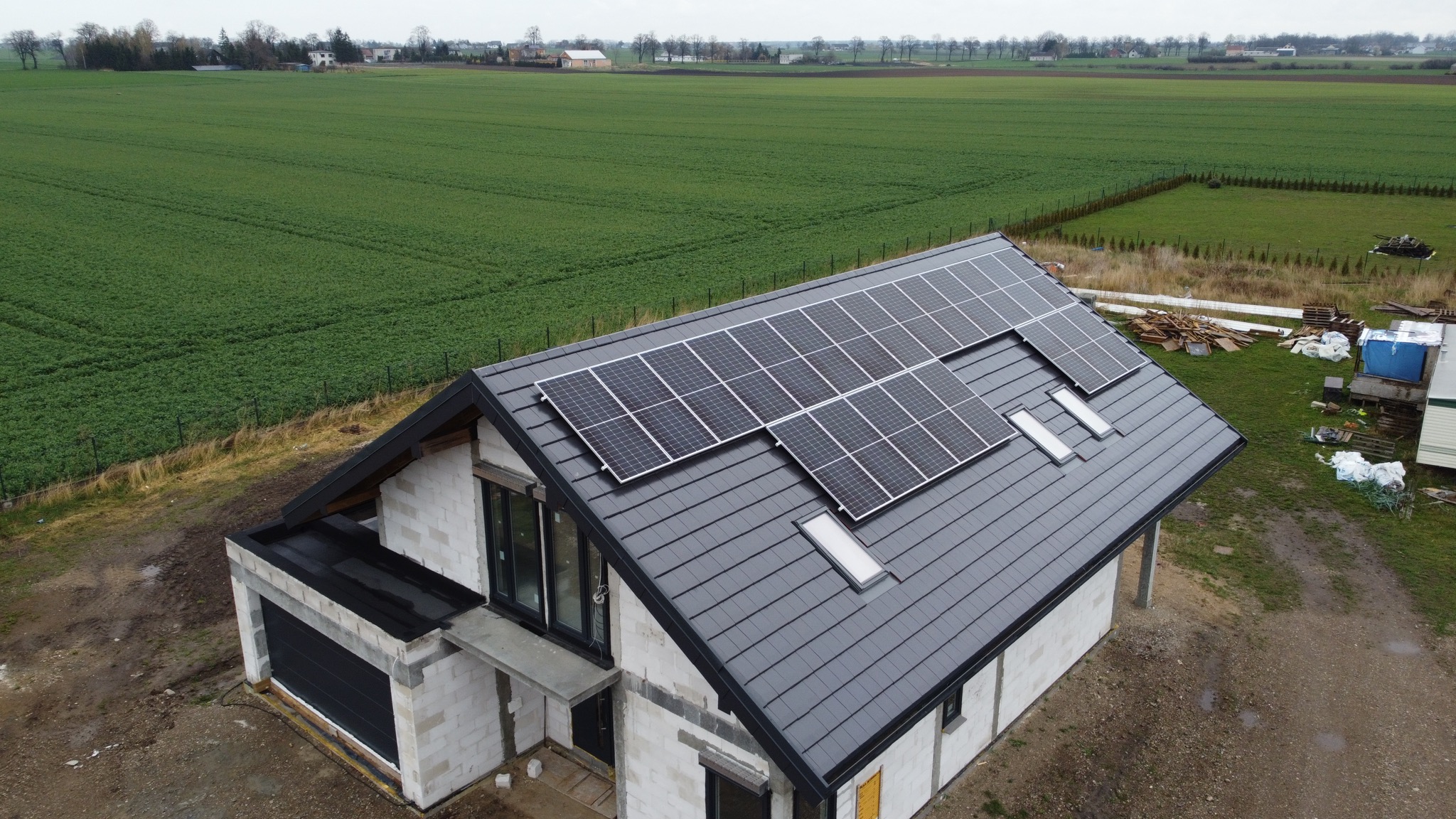 Dom jednorodzinny w stanie surowym otwartym z panelami fotowoltaicznymi na dachu, widok z lotu ptaka na tle zielonego pola.