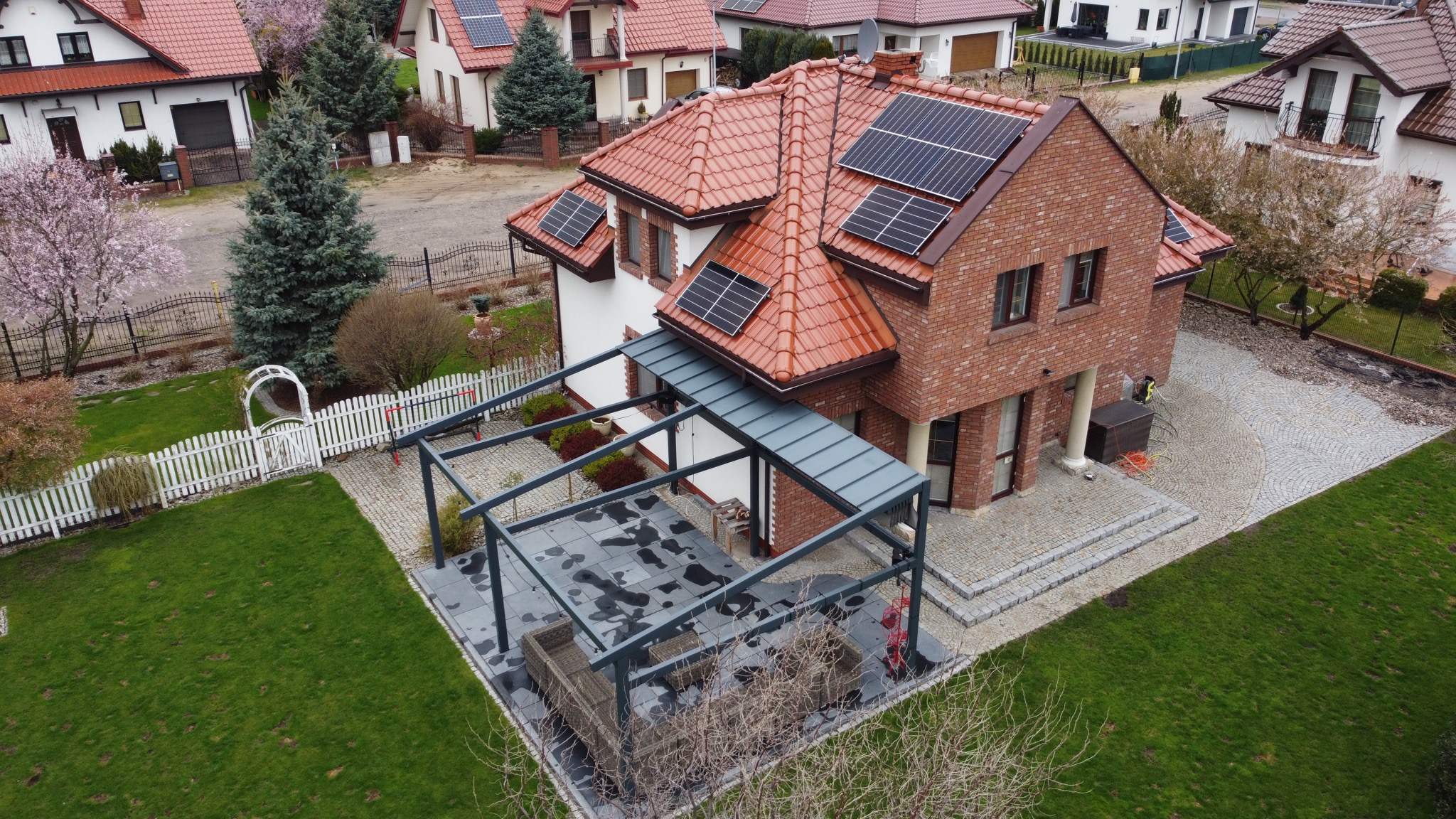 Widok z góry na dom z czerwoną dachówką i panelami słonecznymi, z dobudowaną pergolą nad tarasem, otoczony zielonym trawnikiem i brukowanym podjazdem.