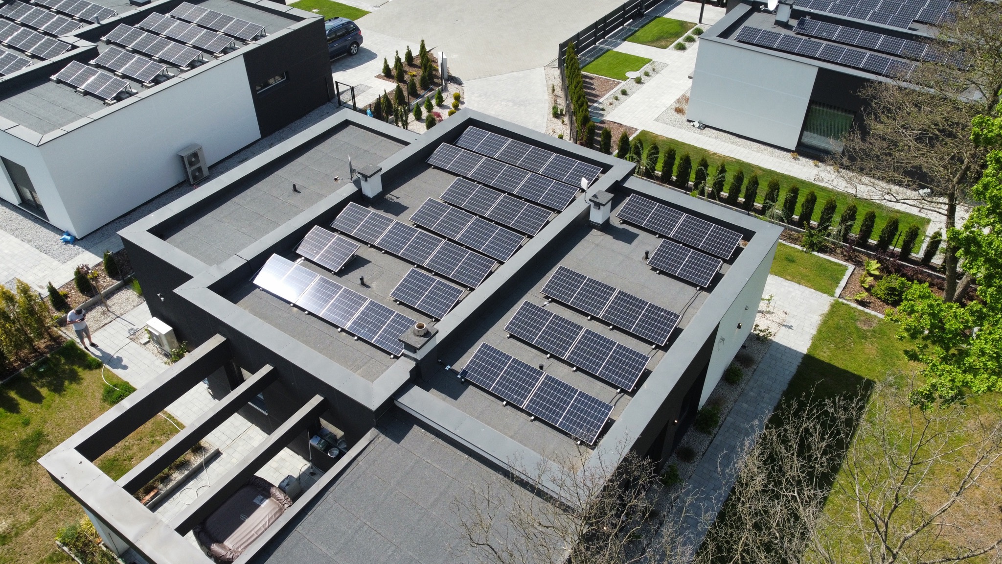 Widok z góry na nowoczesne budynki z panelami słonecznymi na dachu, schludny ogród i osoba spacerująca po posesji.