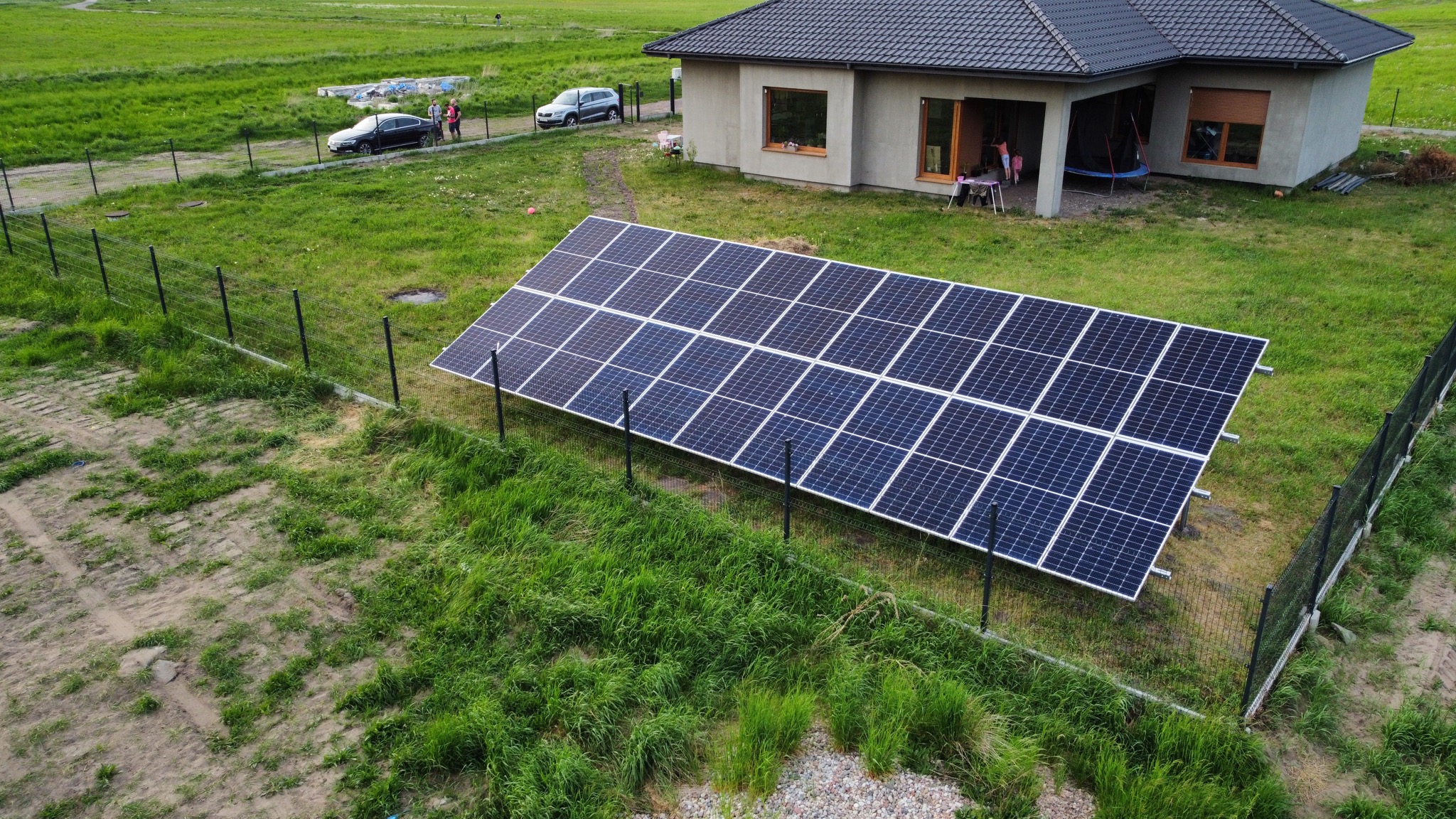 Panel słoneczny na trawniku przed domem jednorodzinnym, widok z góry, czarne ogrodzenie siatkowe w tle.