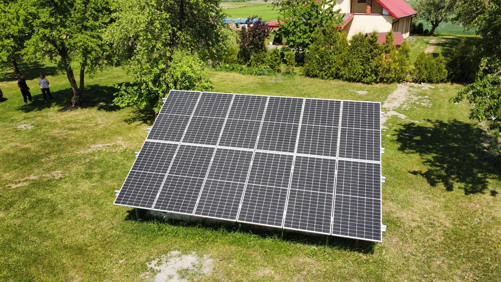 Instalacja fotowoltaiczna na trawniku, widok z góry, w tle dom z czerwonym dachem i drzewa.