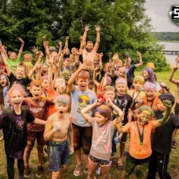 Grupa rozradowanych dzieci z pomalowanymi twarzami i ubraniami, z podniesionymi rękami, świętuje na tle jeziora i lasu, z widocznym logo Survival Race Camp.