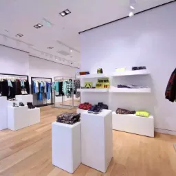 Jasne wnętrze butiku odzieżowego z drewnianą podłogą, białymi ścianami i minimalistycznymi ekspozytorami ubrań i obuwia, oświetlone punktowymi lampami sufitowymi.