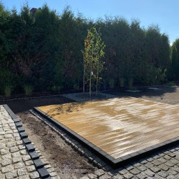 TRZPIOT COMPANY Aleksandra Trzpiot - Platforma z deski kompozytowej z czarnym obramowaniem, obok ścieżka z kostki granitowej w odcieniach szarości z czarnym obrzeżem, w tle rząd wysokich, zielonych tuj i świeżo posadzona trawa...