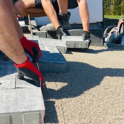 TRZPIOT COMPANY Aleksandra Trzpiot - Układanie szarej kostki brukowej na podsypce z kruszywa, z użyciem linki traserskiej i rękawic ochronnych, widoczna przecinarka do kostki brukowej w tle.