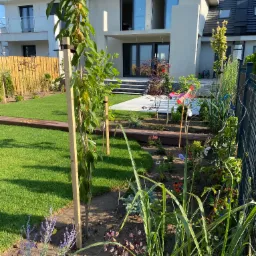 Nowoczesny ogród z trawnikiem, drewnianym chodnikiem, nasadzeniami kwiatów i młodymi drzewkami podpartymi drewnianymi palikami na tle dwukondygnacyjnego domu.