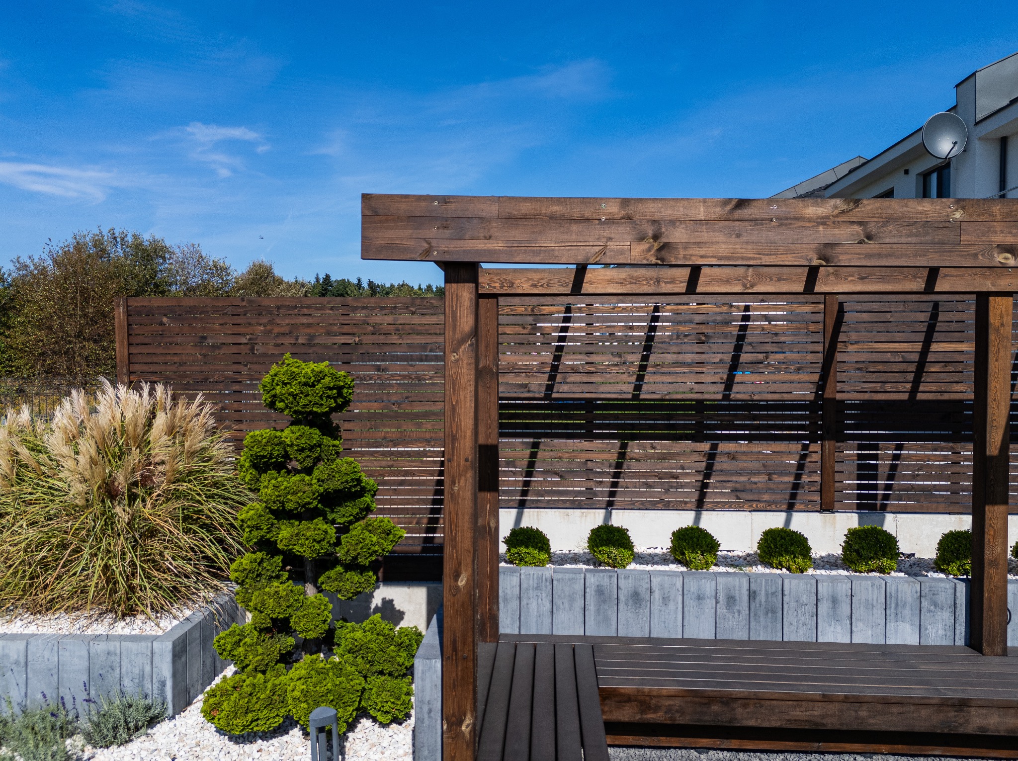 Nowoczesny ogród z drewnianą pergolą, nasadzeniami w donicach z szarej kostki, ozdobnymi trawami i formowanymi krzewami, uzupełniony drewnianą ławką i panelem ogrodzeniowym.