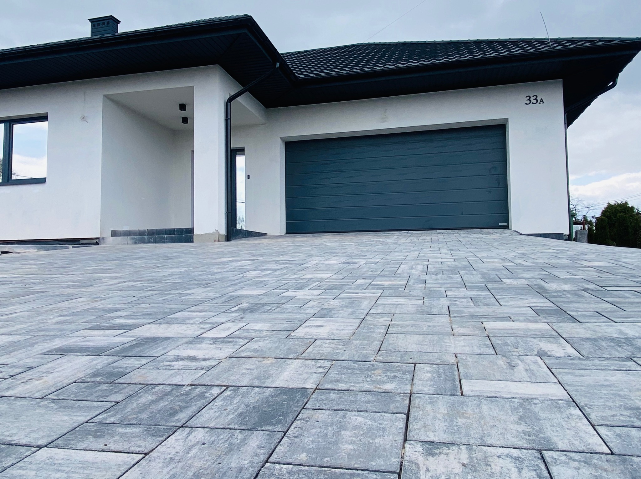 Nowoczesny podjazd z szarej kostki brukowej ułożonej przed białym domem z grafitowym dachem i bramą garażową, widok z niskiej perspektywy.