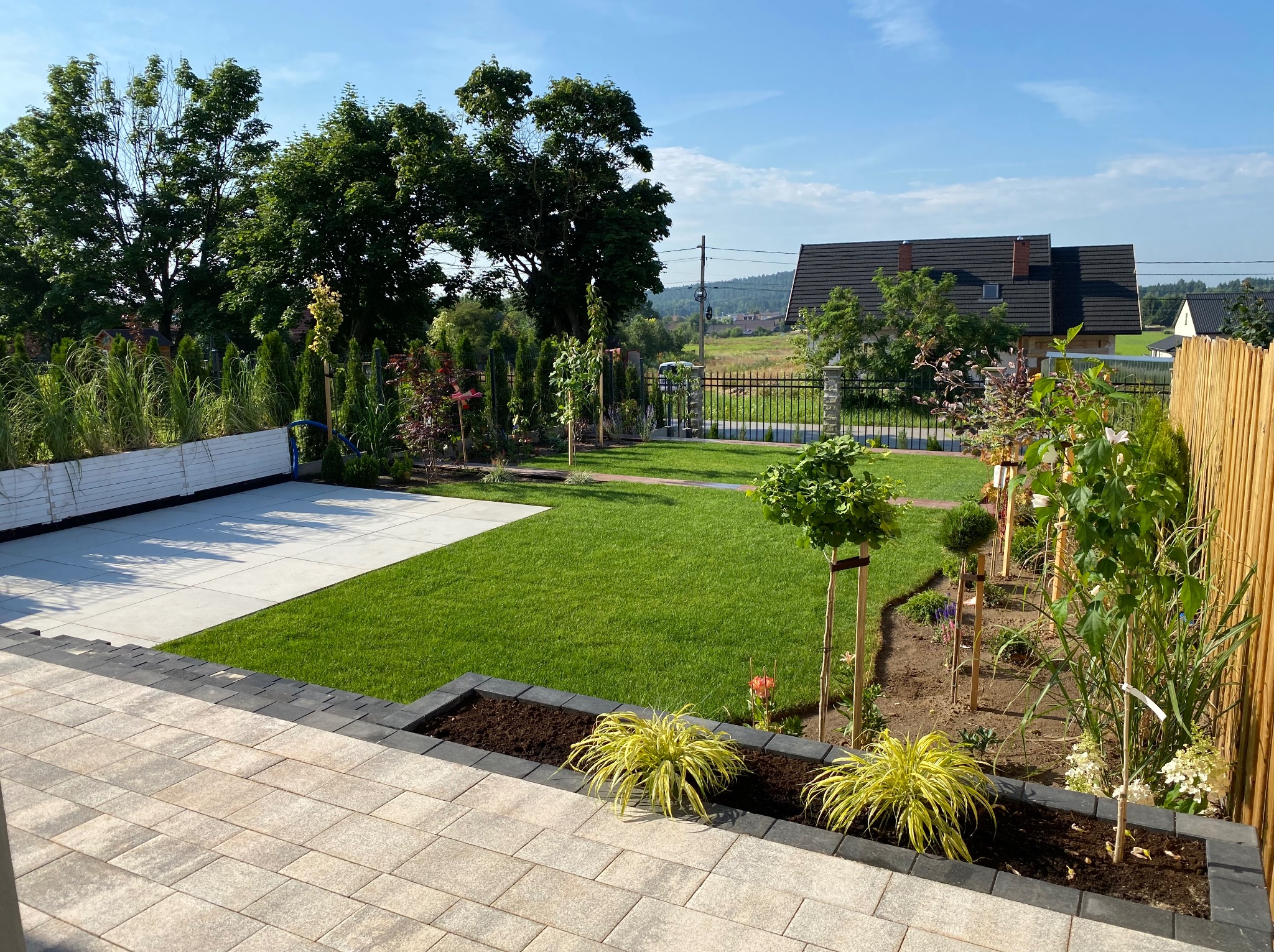Kompleksowo zrealizowany ogród w Górnie z tarasem, trawnikiem z rolki, rabatami kwiatowymi obłożonymi kostką oraz fragmentem nawierzchni z jasnych płyt.