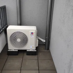 Zewnętrzna jednostka klimatyzacji Gree Inverter zamontowana na balkonie, widoczne rury odprowadzające skropliny i czarne podkładki antywibracyjne.