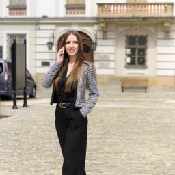 Kobieta w czarnym kombinezonie i marynarce w pepitkę rozmawia przez telefon na tle klasycznego budynku z jasną fasadą i brukowanym placem.