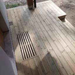Nowy taras z jasnego drewna z wbudowanymi kratkami odpływowymi i pojedynczym stopniem, częściowo zacieniony.