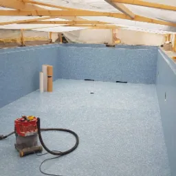 Wnętrze basenu w trakcie wykańczania, wyłożone jasnoniebieską mozaiką, widoczny odkurzacz przemysłowy, rolki materiału i narzędzia, konstrukcja drewniana dachu w tle.