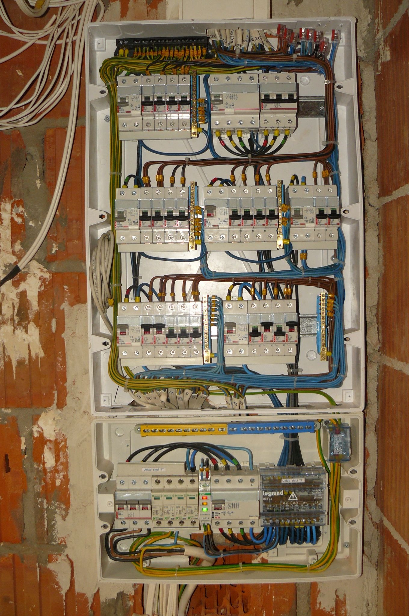 Otwarta skrzynka elektryczna z licznymi bezpiecznikami i przewodami, zamontowana na surowej ścianie z cegły, widoczne oznaczenia 'Legrand' i 'Uklad sieci TT'.