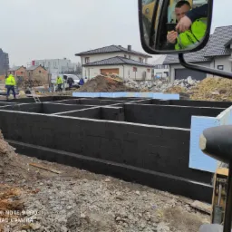 Przygotowany szalunek pod fundamenty domu jednorodzinnego, zabezpieczony hydroizolacją i ocieplony styropianem, widok z kabiny maszyny budowlanej, w tle pracownicy budowlani i zabudowa mieszkalna.