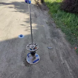 Urządzenie do badania zagęszczenia gruntu na szutrowej drodze, widoczny ślad koła i luźny kamień.