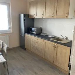 Jasna kuchnia w mieszkaniu z drewnopodobnymi płytkami podłogowymi, beżowymi szafkami i szarym blatem, uzupełniona o srebrną lodówkę i mikrofalówkę.