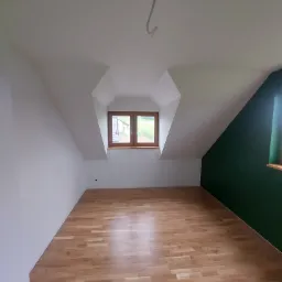 Poddasze po remoncie z drewnianą podłogą, oknem w szczycie dachu i ścianą pomalowaną na zielono, widoczny kabel wystający z sufitu.