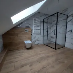 Nowoczesna łazienka na poddaszu z drewnopodobną podłogą, marmurowymi płytkami ściennymi, kabiną prysznicową z czarnymi ramami i podwieszaną toaletą.