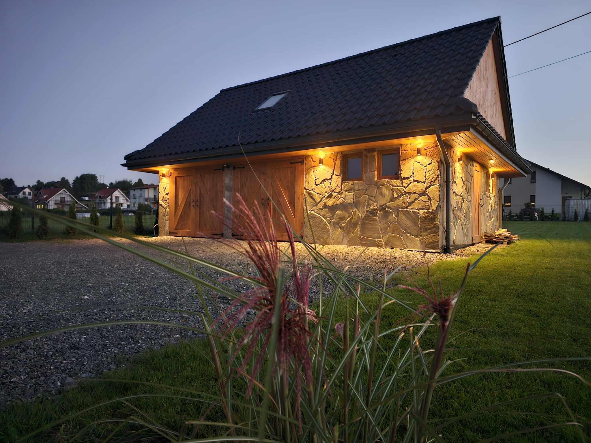 Kamienny budynek gospodarczy z drewnianymi wrotami garażowymi, oświetlony ciepłym światłem, na tle wieczornego nieba i zieleni. Detal: ozdobna trawa na pierwszym planie.