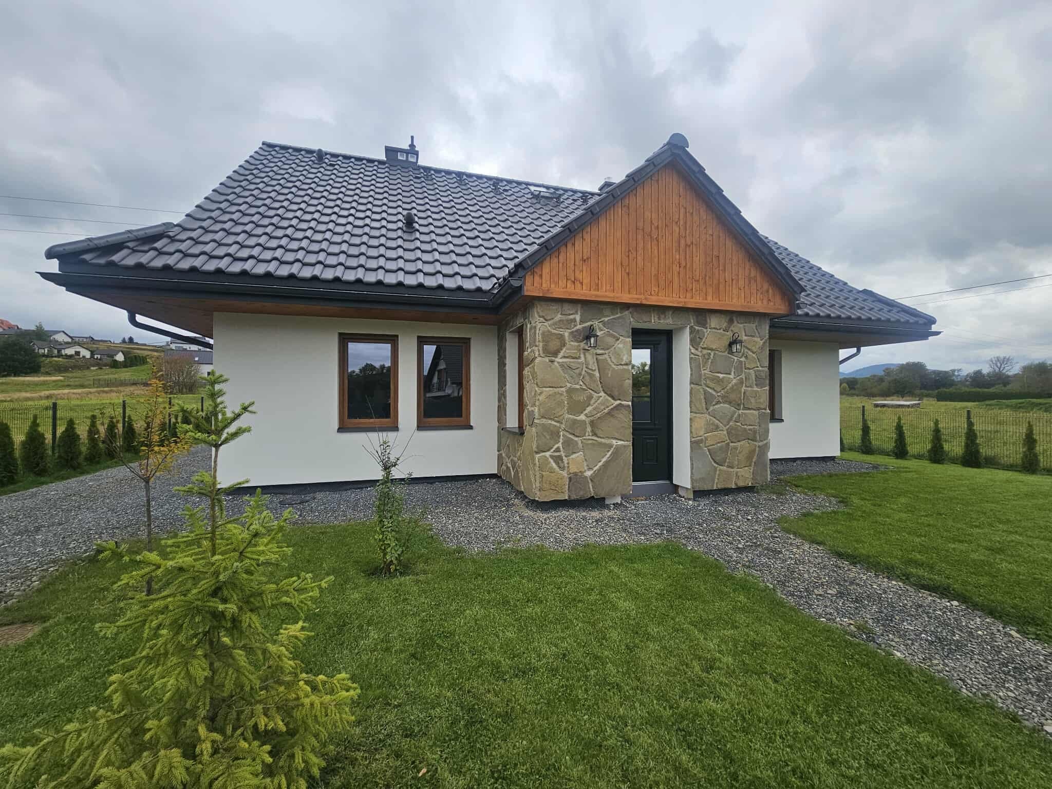 Nowoczesny dom jednorodzinny z elewacją z kamienia i drewnianym szczytem, otoczony zielonym trawnikiem i ścieżką z kamienia. Dach pokryty ciemną dachówką.