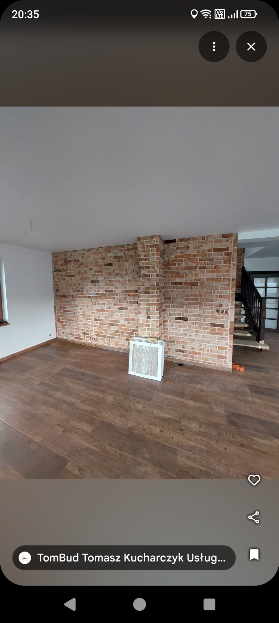 Wykończone wnętrze z ceglaną ścianą i drewnopodobną podłogą. Widoczne schody z czarną balustradą. W rogu pomieszczenia leżą białe panele.