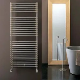 Chromowany drabinkowy grzejnik łazienkowy z mosiężnymi detalami, wiszący na ciemnobrązowej ścianie, obok lustra w złotej ramie i wolnostojącej baterii umywalkowej.