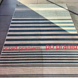 Pasiasty dywan na balkonie z tekstem 'przed praniem' i 'po praniu' naniesionym na zdjęcie, dolna krawędź częściowo zasłonięta przez niebieskie urządzenie czyszczące.