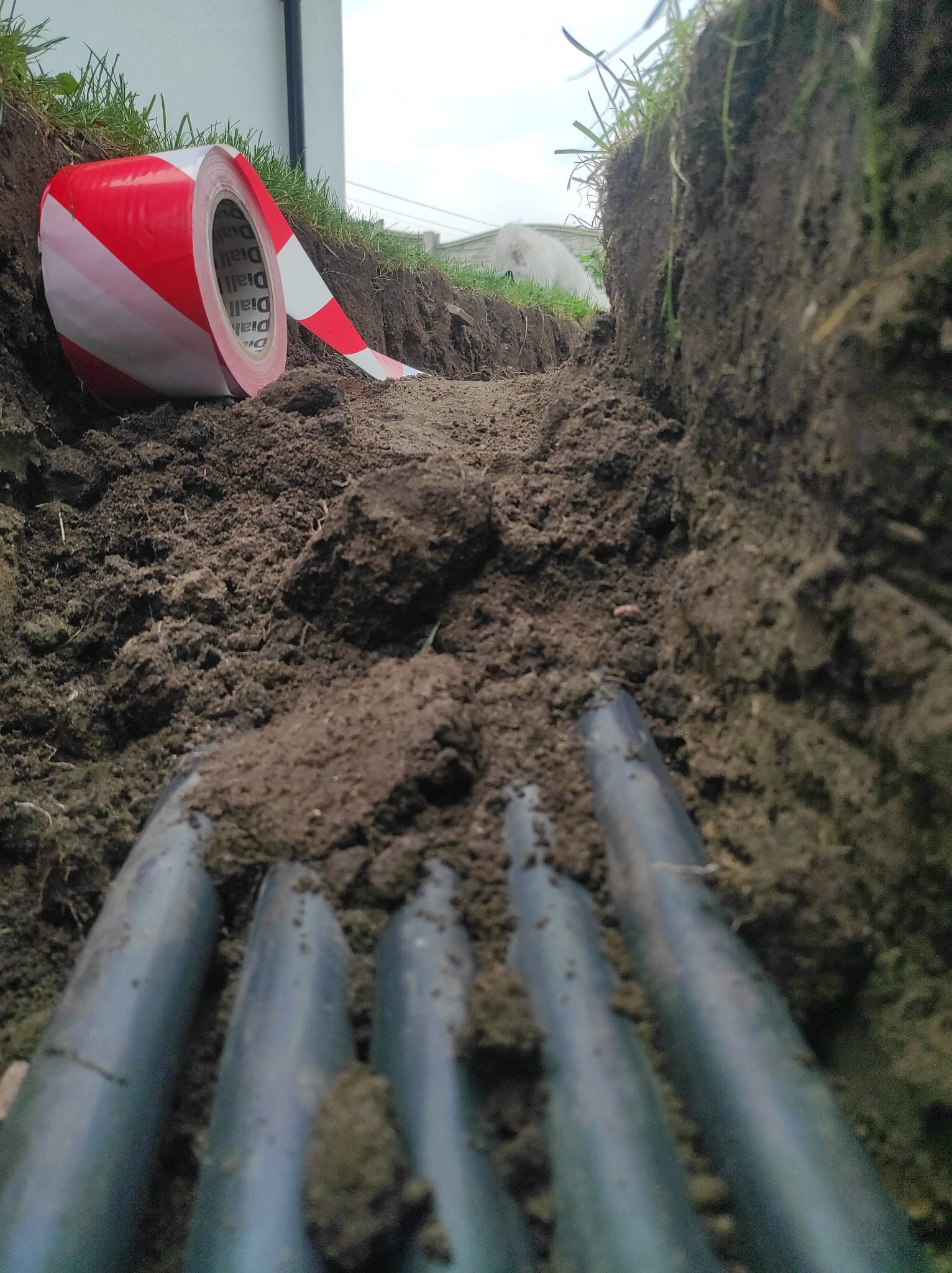 Wykop w ziemi z ułożonymi rurami instalacyjnymi, zabezpieczony taśmą ostrzegawczą z rolki.
